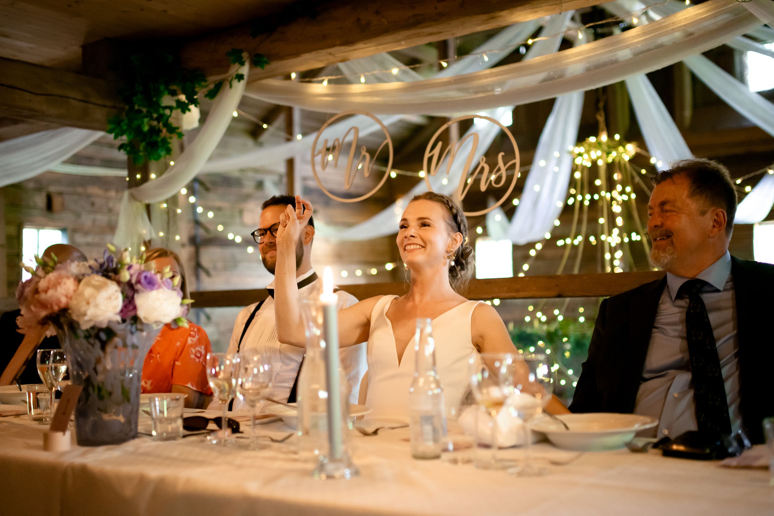 Wedding reception dinner scene with decorative lighting and white fabric draping overhead.