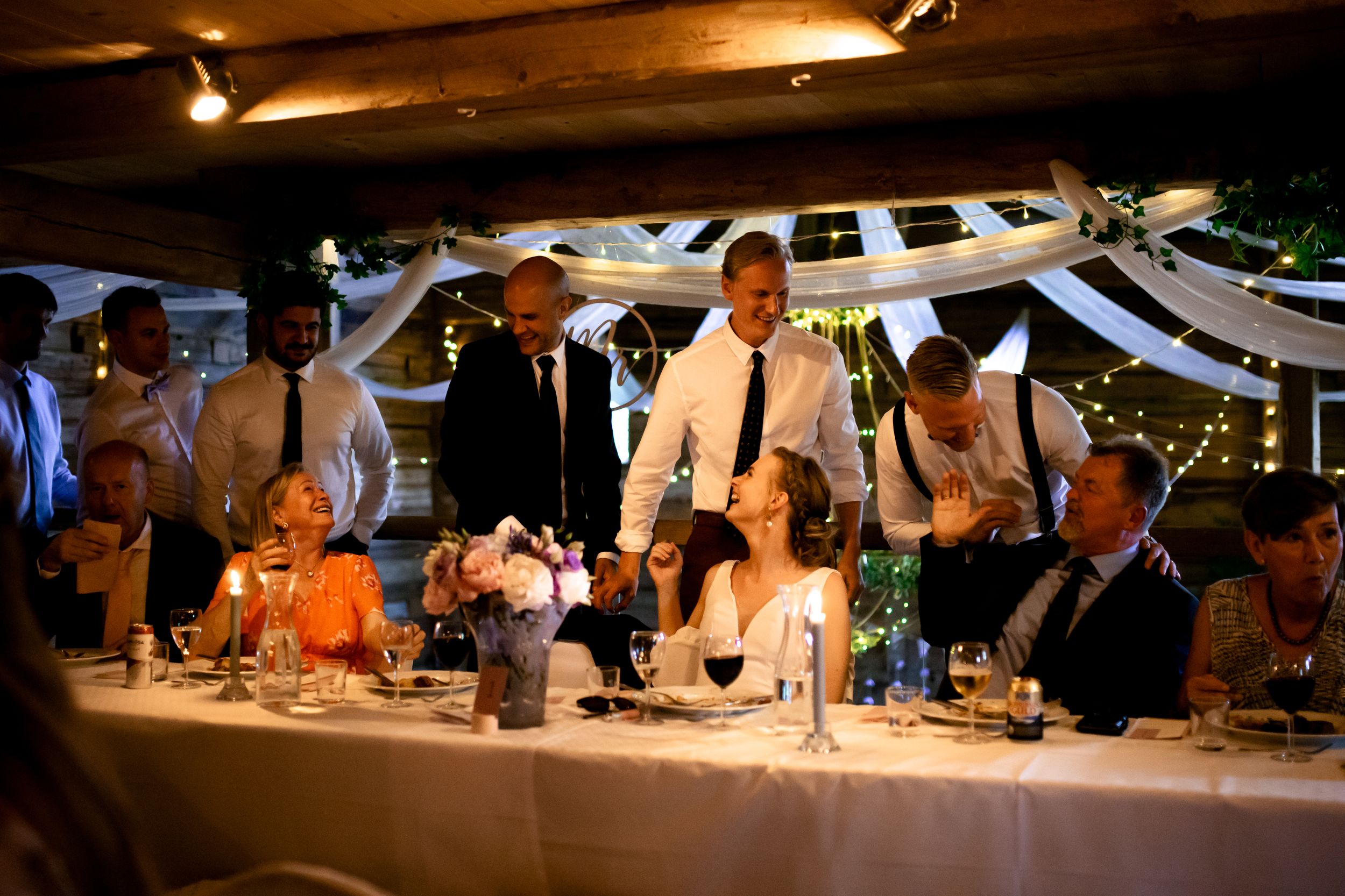Wedding reception celebration in rustic venue with hanging lights and white decorations.