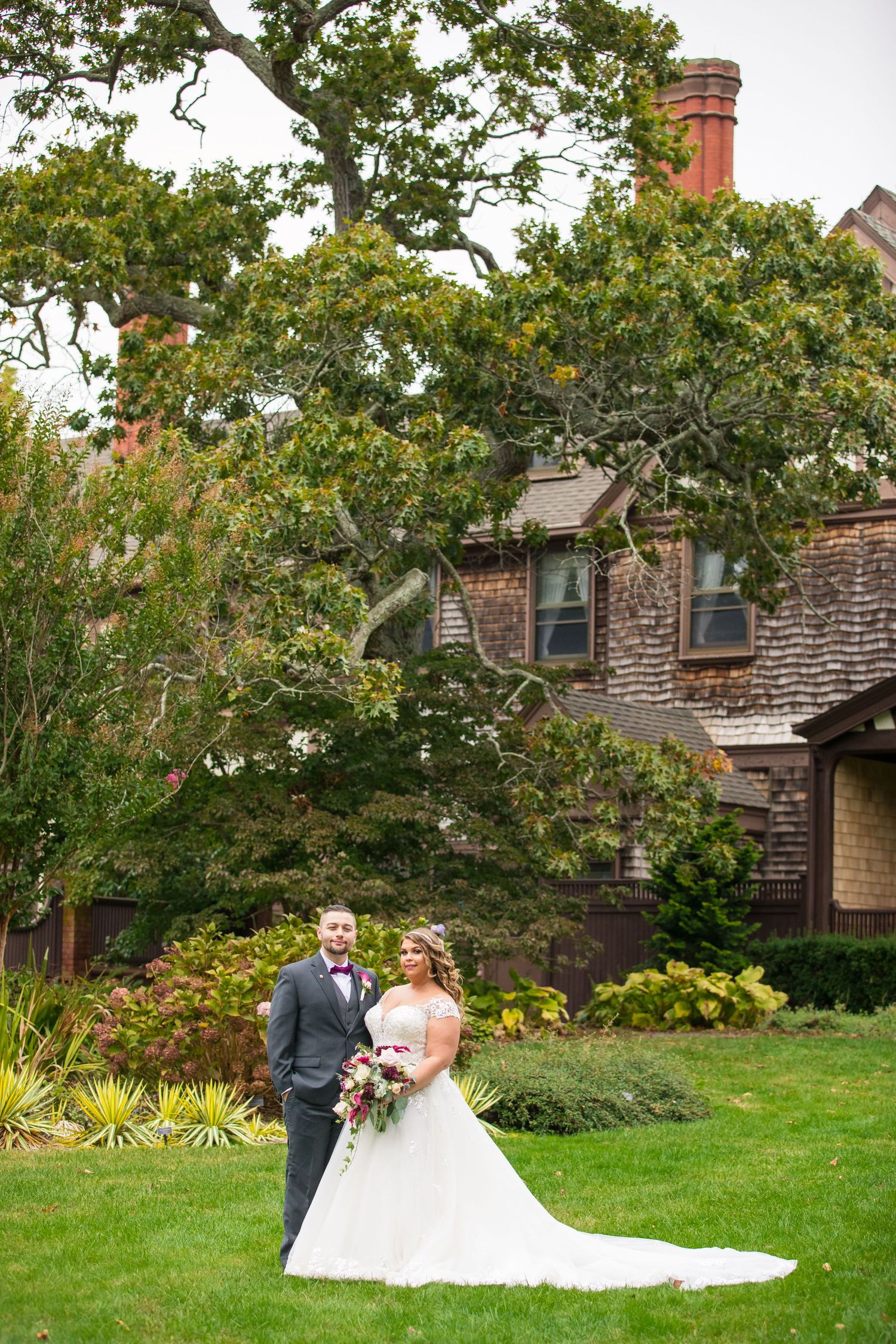 Wedding portrait in front of a historic brick house with green gardens and trees.