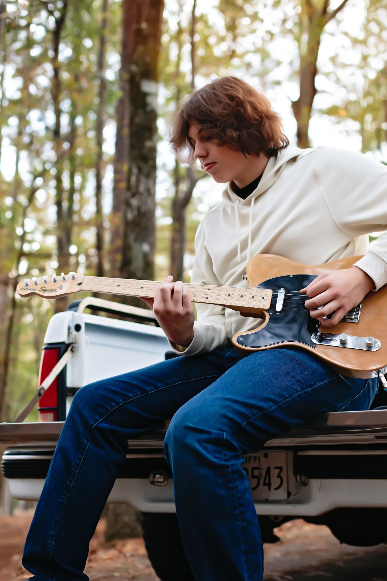 A person in a white hoodie plays bass guitar while sitting on the back of a pickup truck.