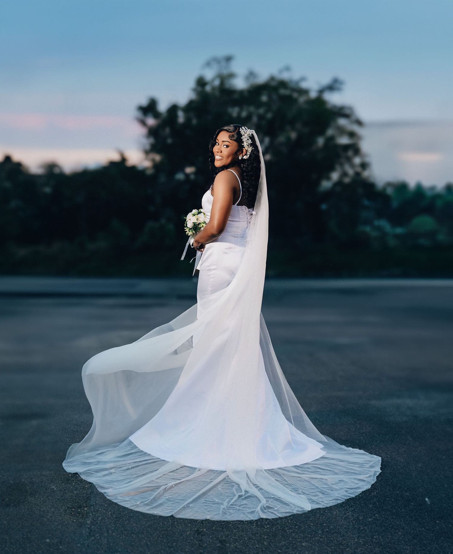 Bride in flowing white wedding gown with long veil holding bouquet at twilight outdoors.