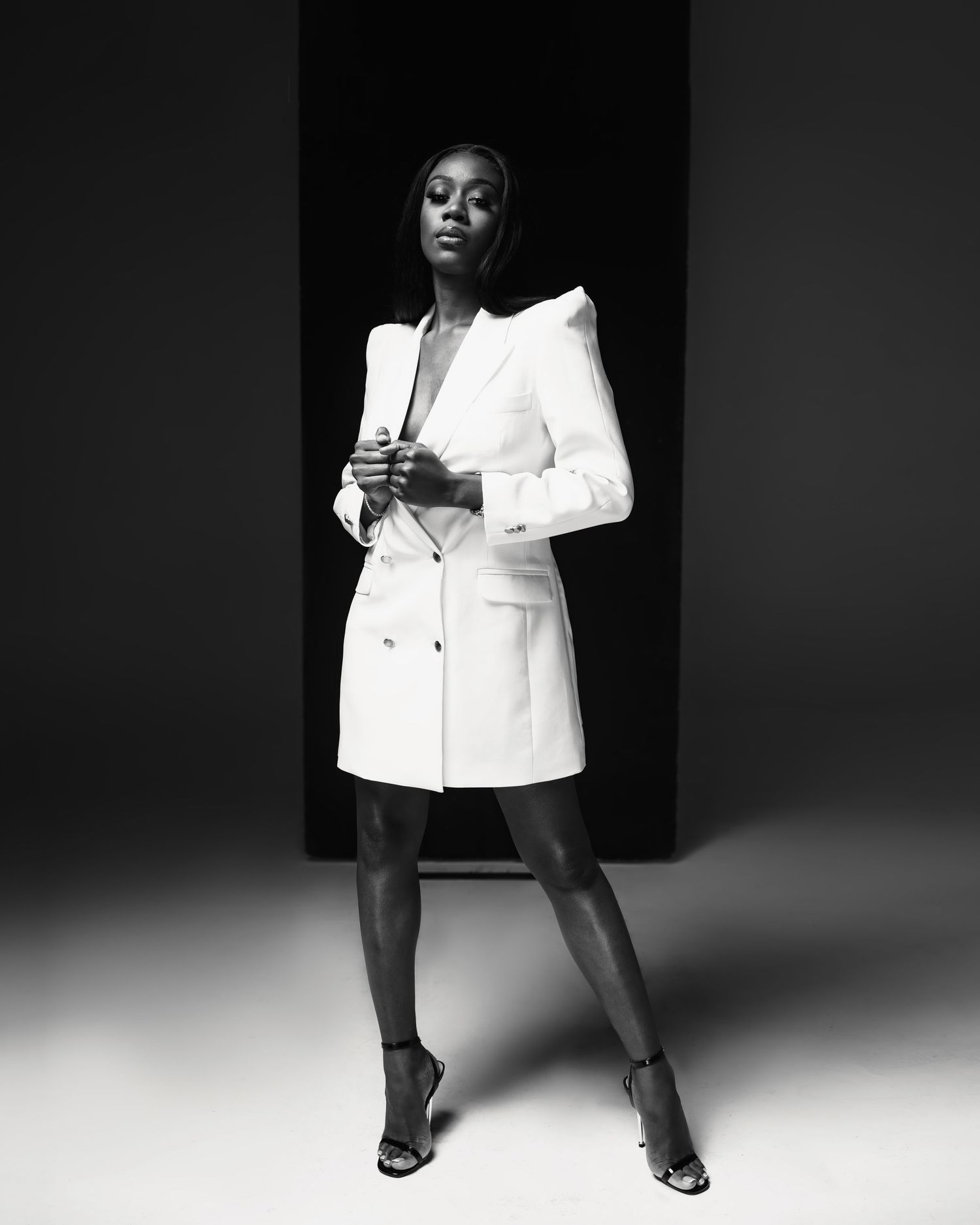 Fashion portrait of model in white blazer dress striking confident pose against dark background.