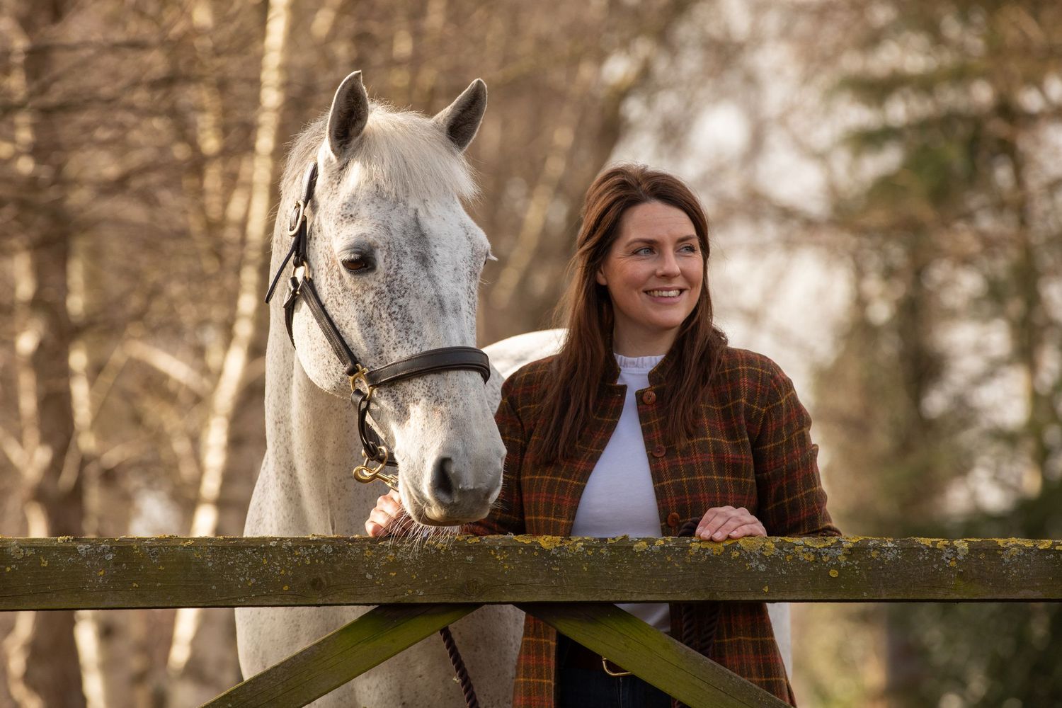 A beautiful white horse poses with someone in a brown jacket at a wooden fence.