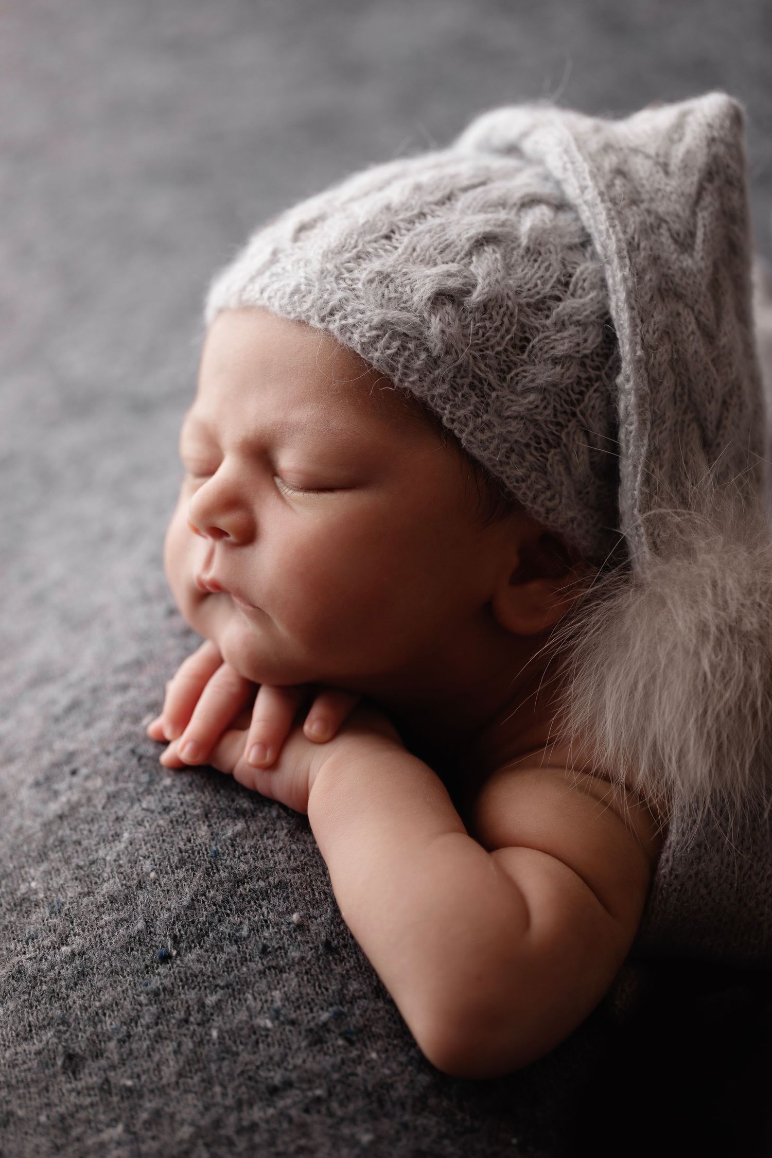 A sleeping newborn wearing a knit cap rests on grey textured material.