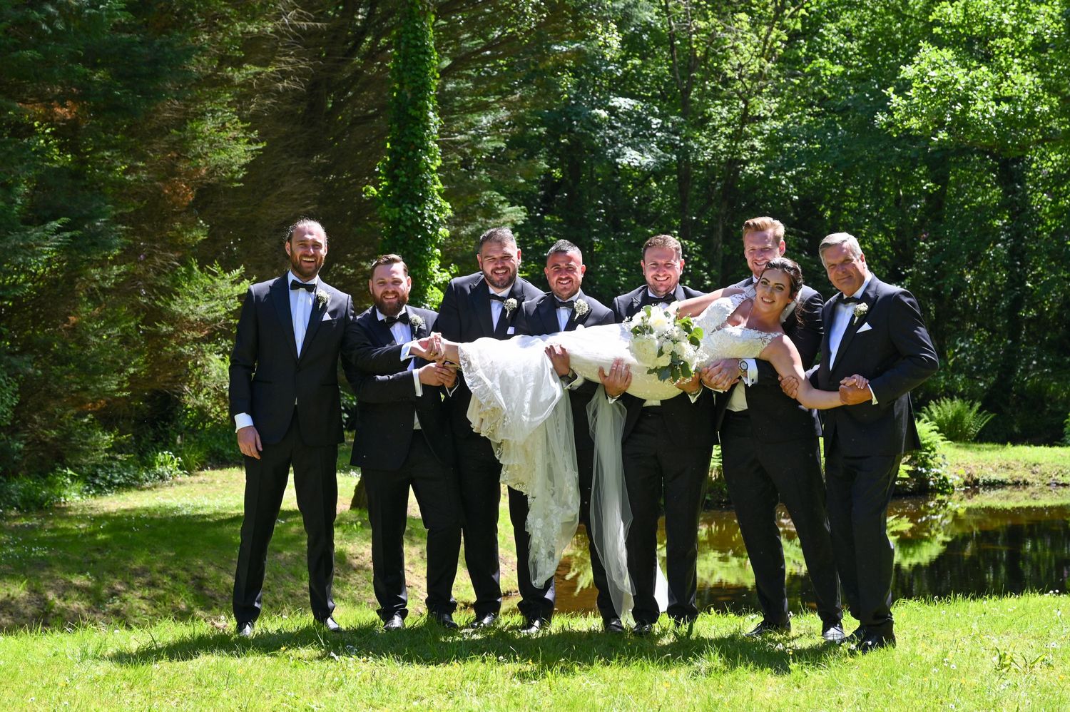 Wedding party members in formal attire pose together while lifting someone horizontally outdoors.