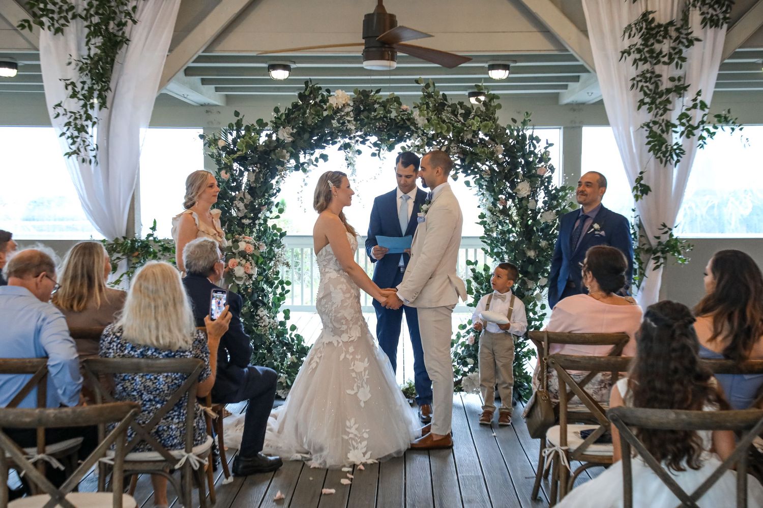 A beautiful outdoor wedding ceremony taking place under a white gazebo decorated with floral arches by the ocean.