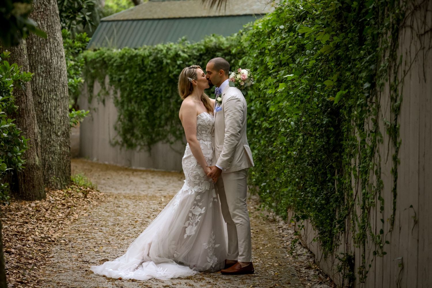 A couple in wedding attire share an intimate moment in a lush garden pathway with ivy-covered walls.