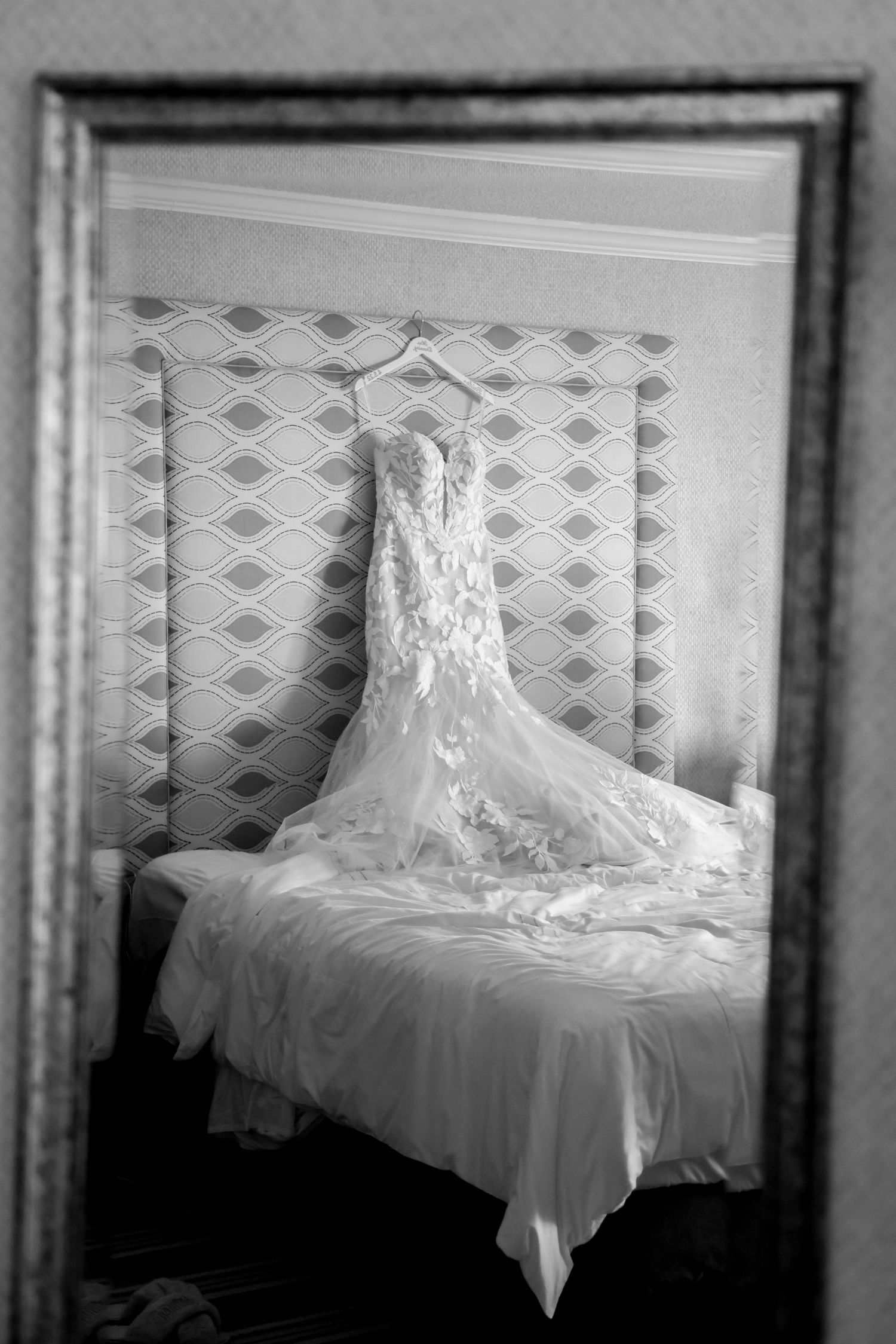 A black and white photograph of an elegant lace wedding dress displayed on a bed against a patterned wall.