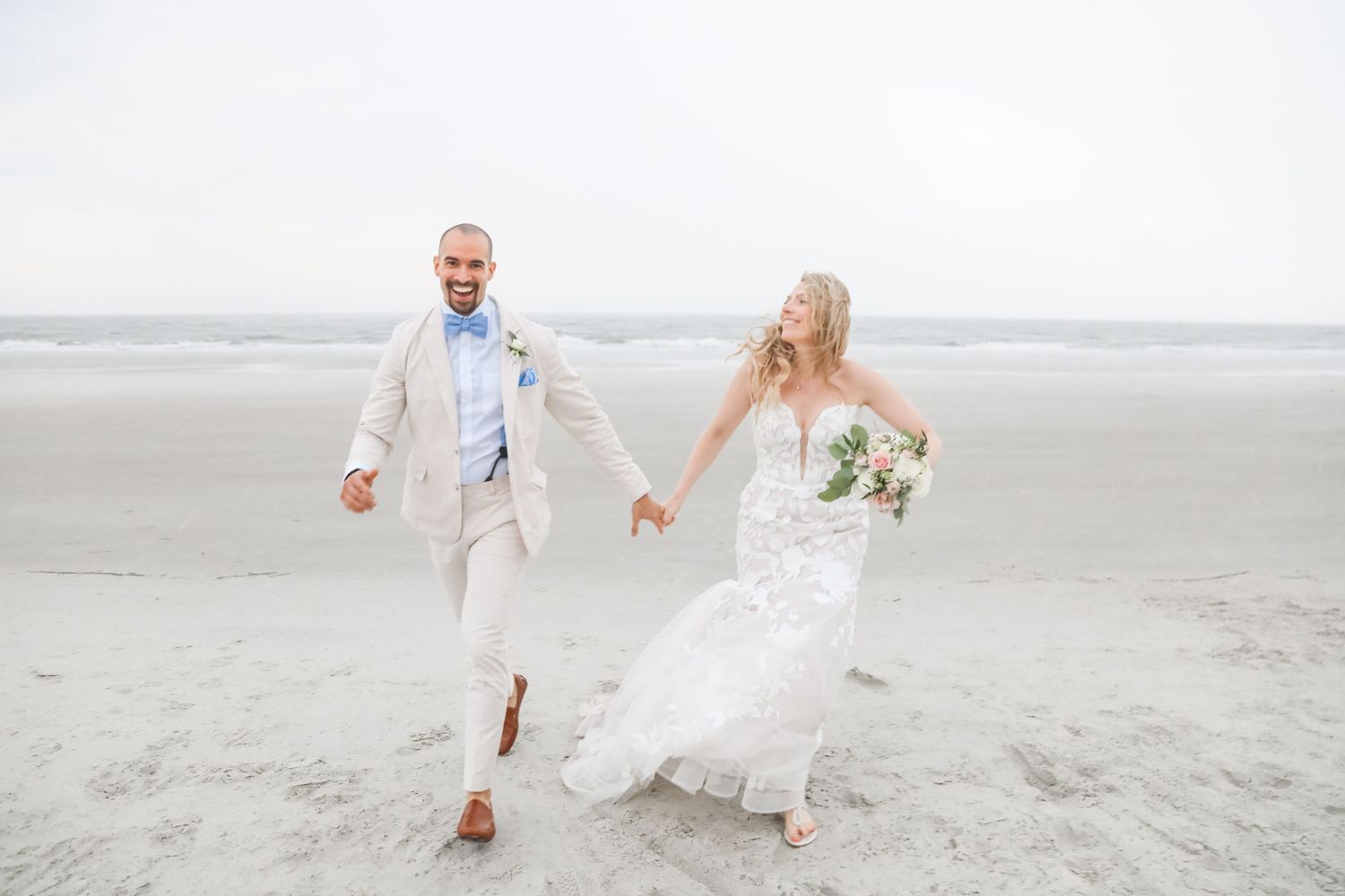 A happy couple runs joyfully along a pristine beach, with the bride in white holding a bouquet on their wedding day.