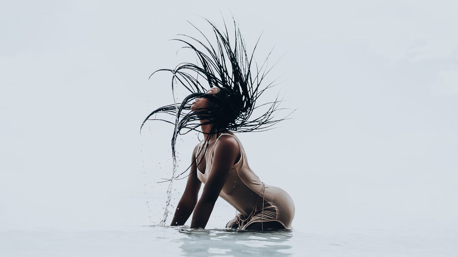 Dramatic shot of a figure flipping their hair back in water against light background.