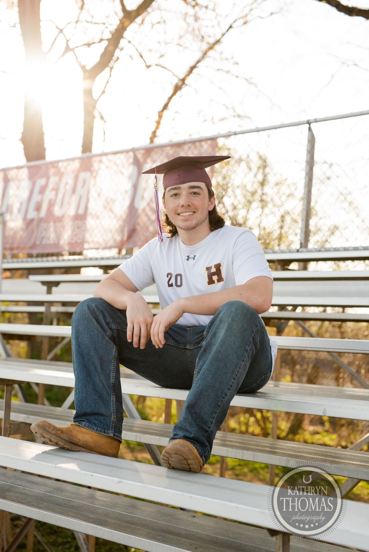 Graduating student in a white jersey with number 20 sits casually on bleachers during outdoor photo session.
