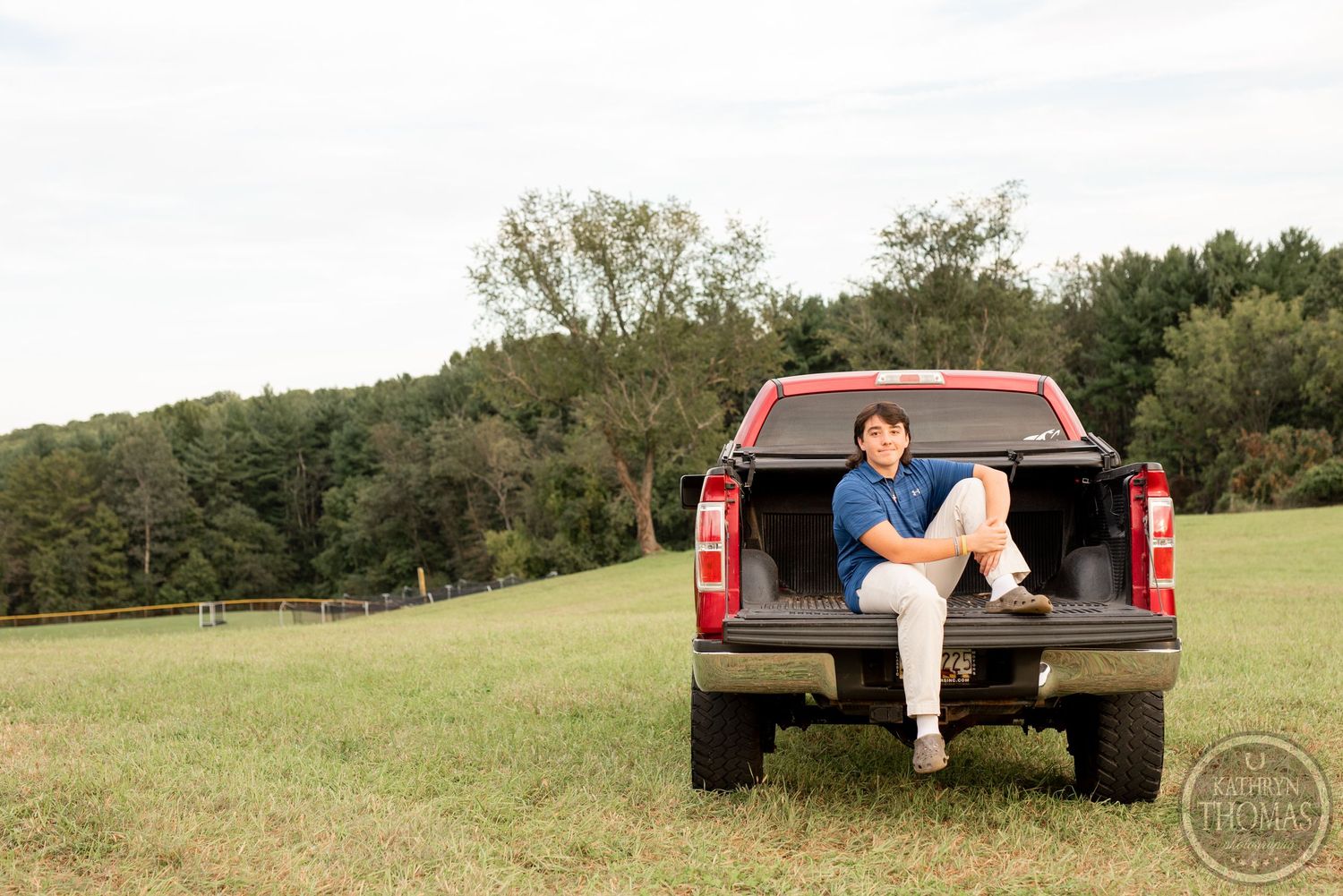 A casual outdoor photo of someone sitting on the tailgate of a red pickup truck in a field.