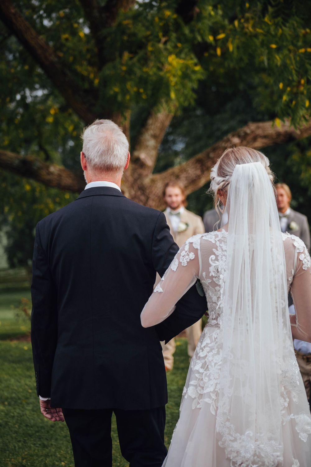 Bride in lace gown and veil walks arm-in-arm with older man in black suit toward outdoor wedding ceremony.