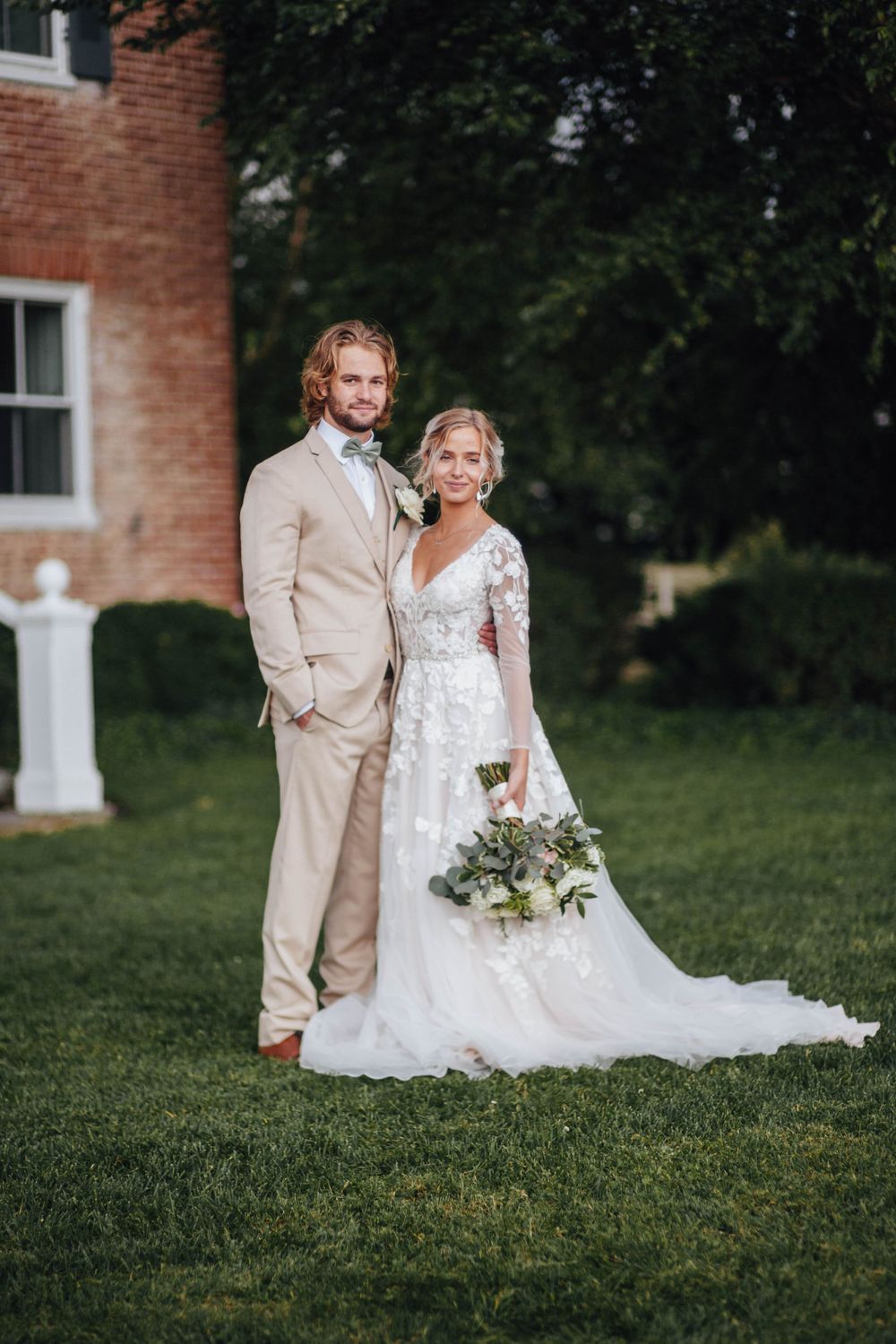 Newlyweds pose on green lawn, groom in tan suit, bride in lace wedding dress holding a floral bouquet.