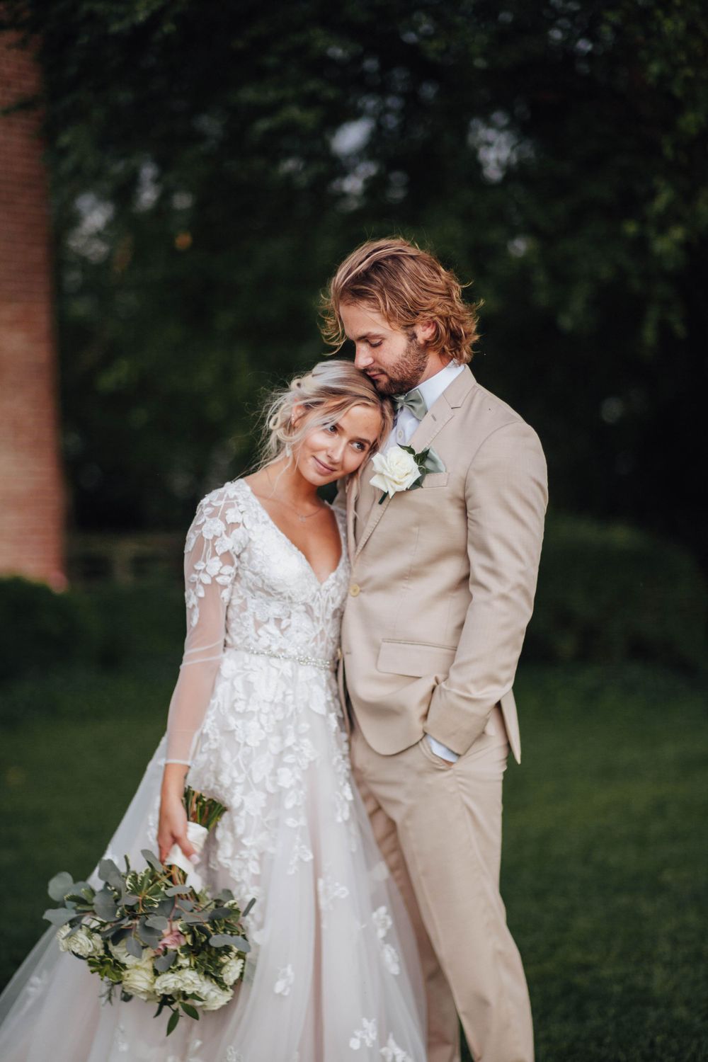 Bride in lace V-neck gown leans on groom in tan suit holding white bouquet in lush green garden setting.