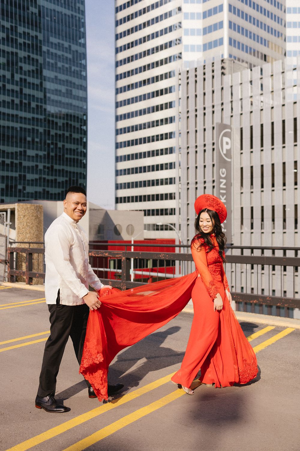 A couple poses playfully with flowing red fabric against an urban backdrop of office buildings.