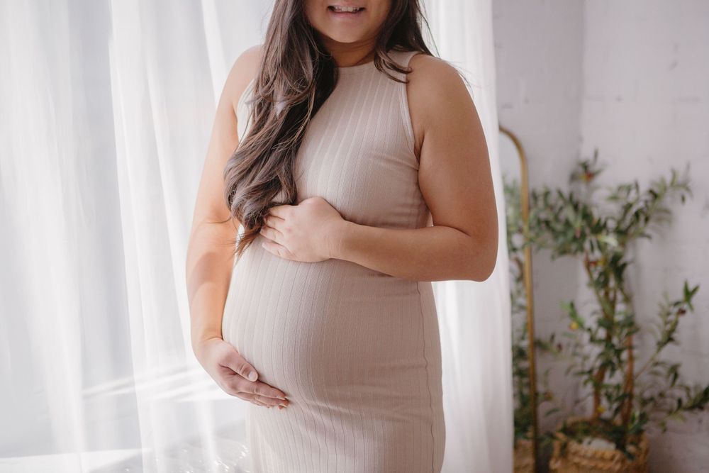 Woman in a beige dress poses confidently against a white backdrop with natural lighting.
