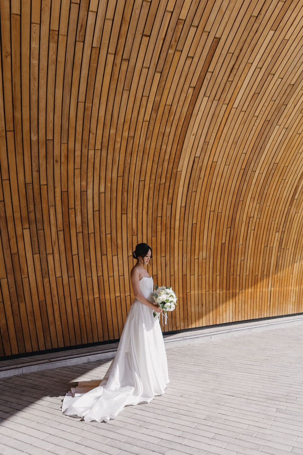 Bride in flowing white dress poses against curved wooden architectural backdrop.