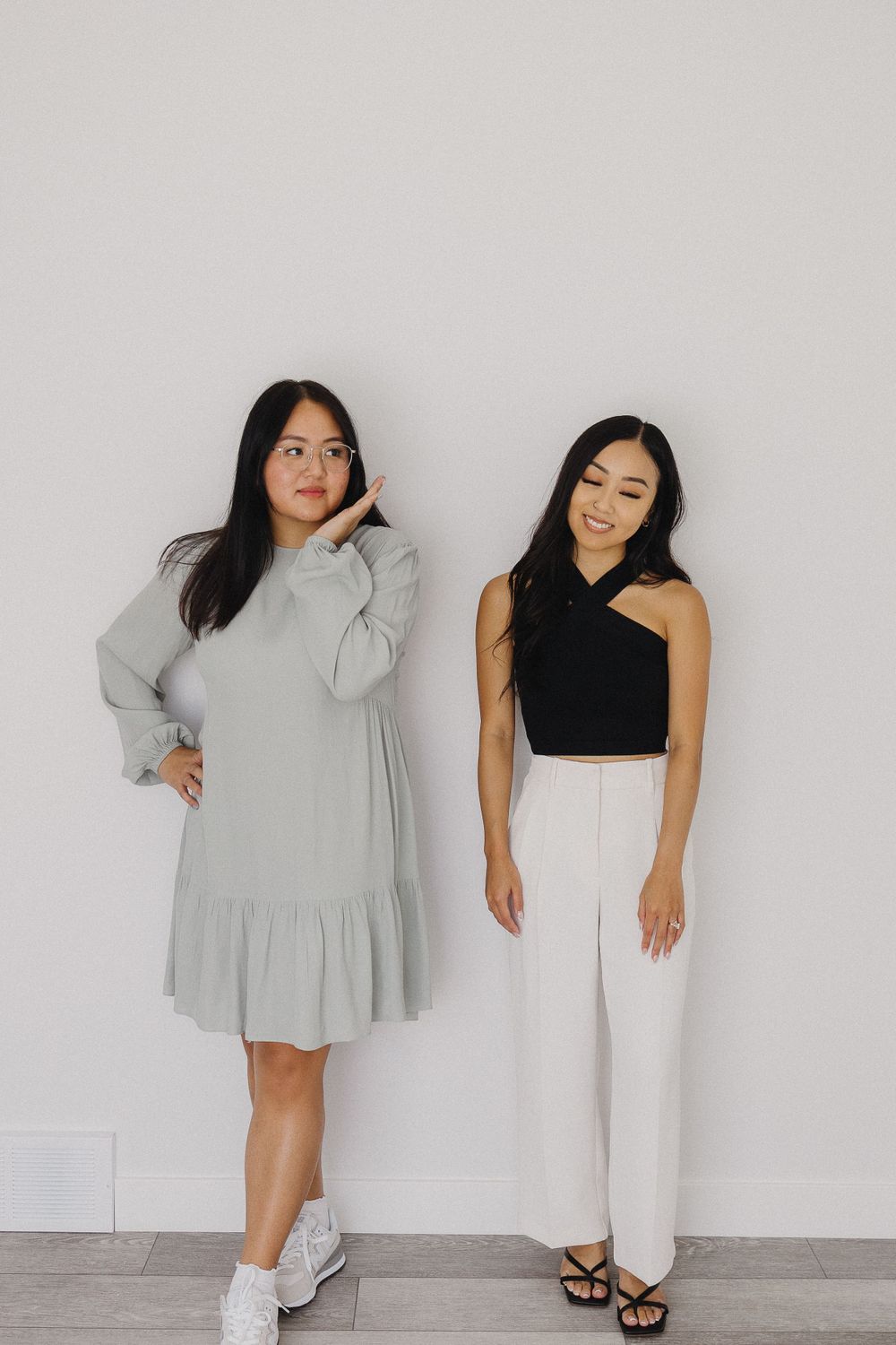 Two friends posing playfully in contrasting outfits against a minimalist grey wall.