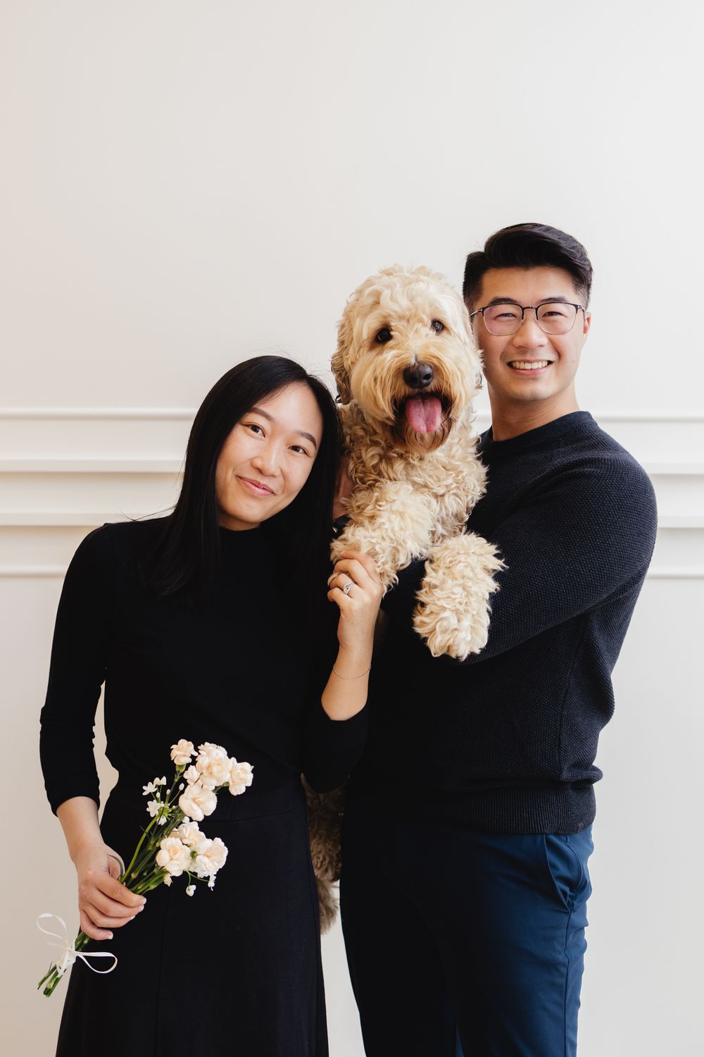 A couple poses with their fluffy goldendoodle dog against a white wall while holding flowers.