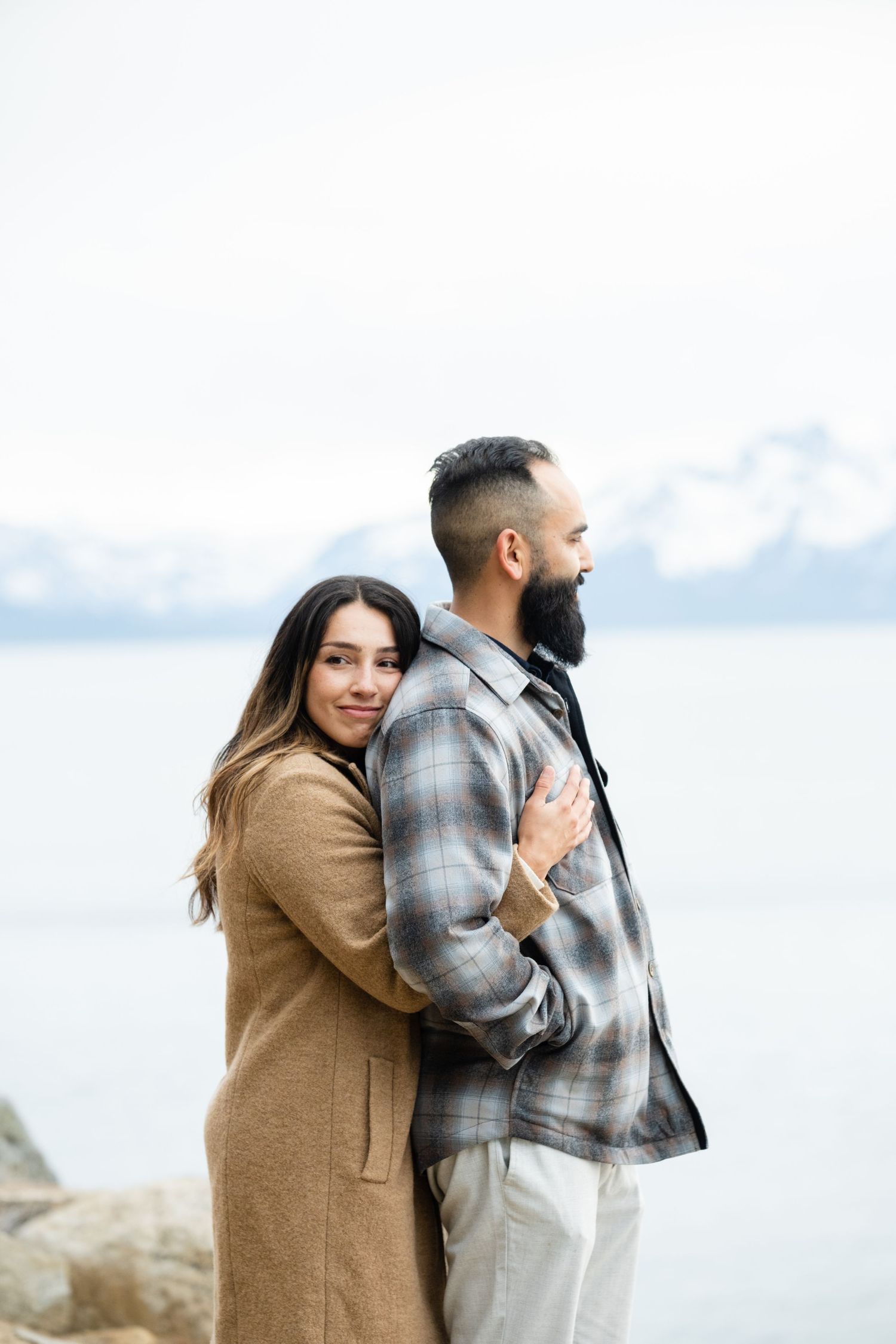 A couple embraces while standing on rocky shoreline overlooking calm water and cloudy sky.