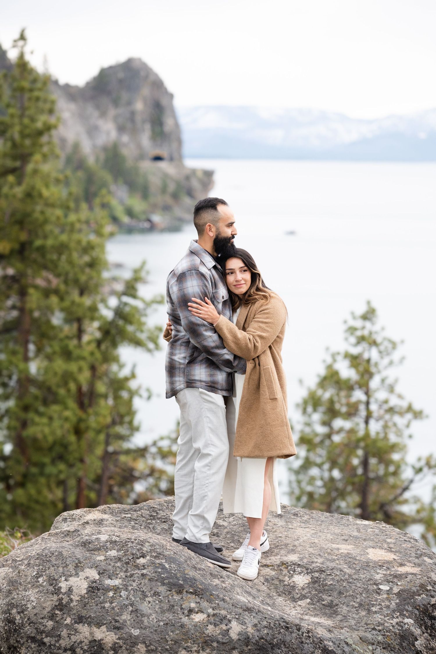 A couple embraces while standing on a large rock overlooking a scenic mountain lake landscape.