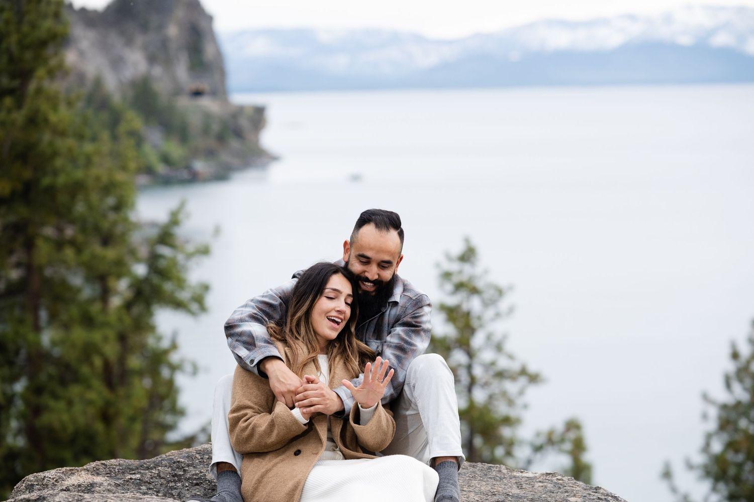 A couple sits lovingly on a cliff edge overlooking a misty lake with mountain backdrop.