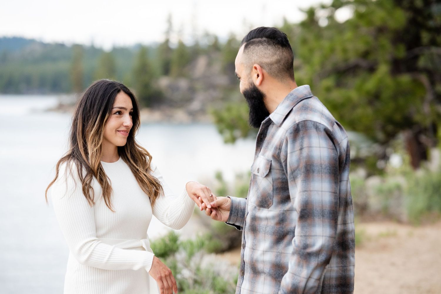 A couple shares a romantic moment by a tranquil lakeside with evergreen trees in the background.
