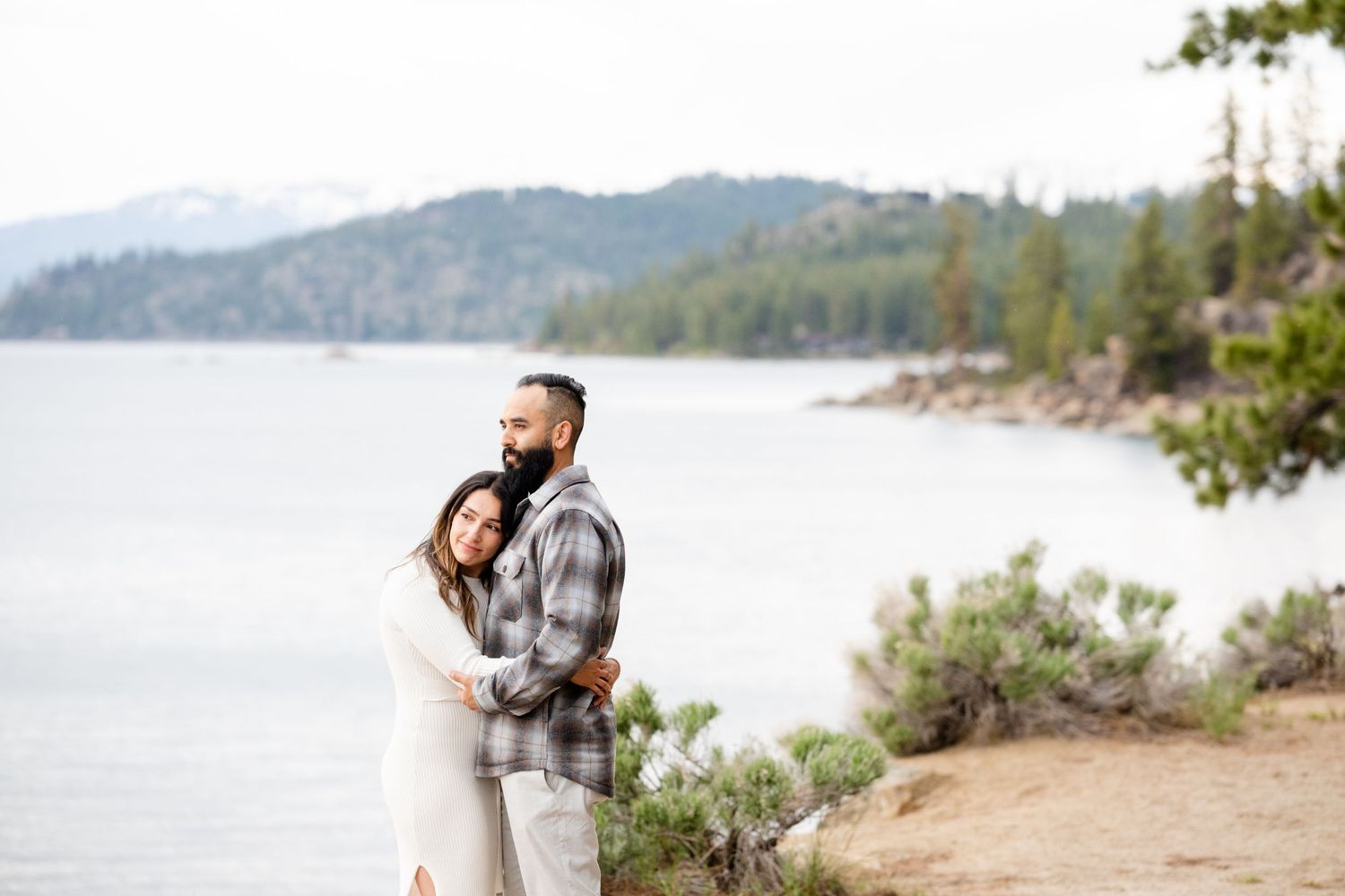 A couple embraces while standing on a rocky shoreline with mountains and a tranquil lake in the background.