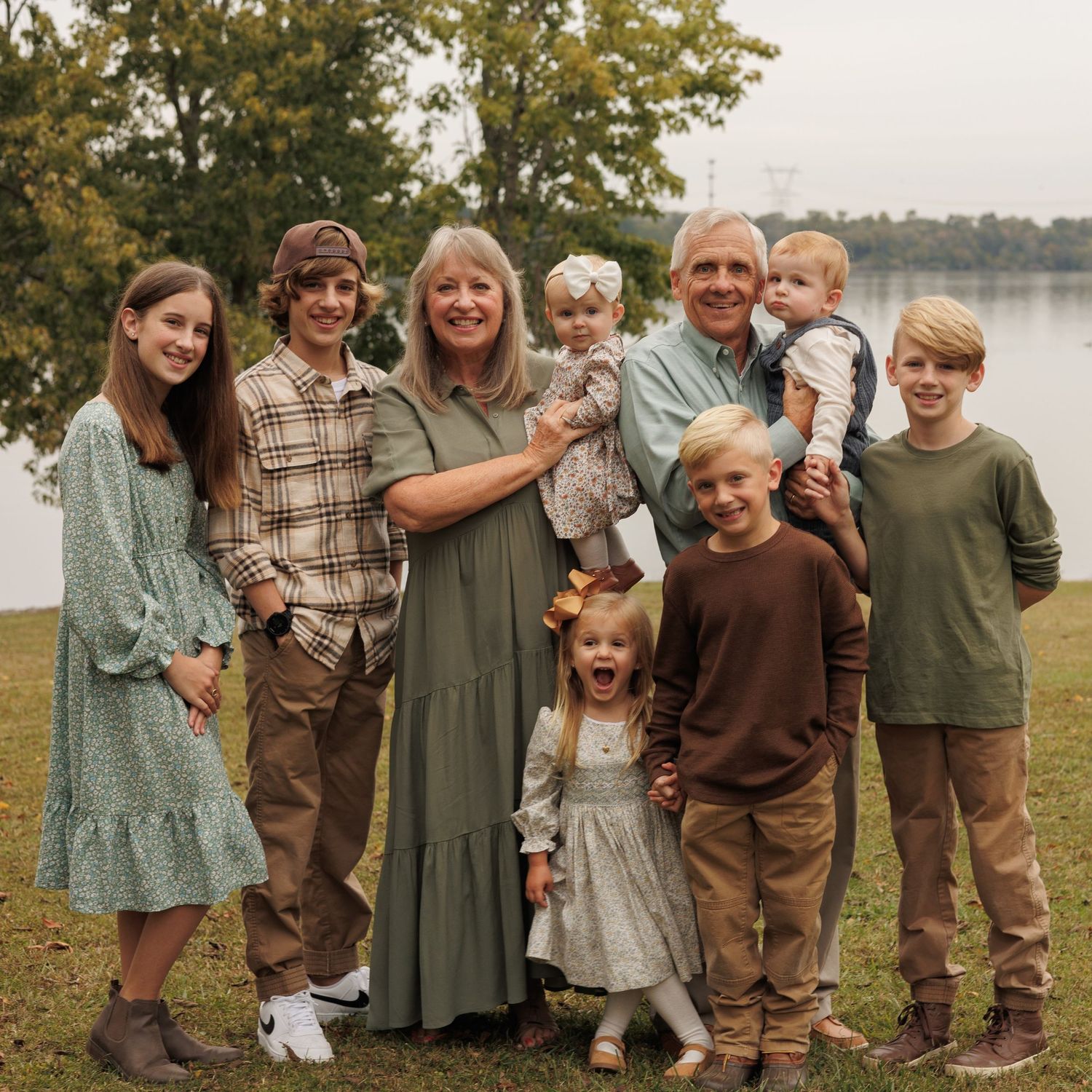 Group portrait outdoors by a lake with everyone dressed in casual earth tones and green dresses.