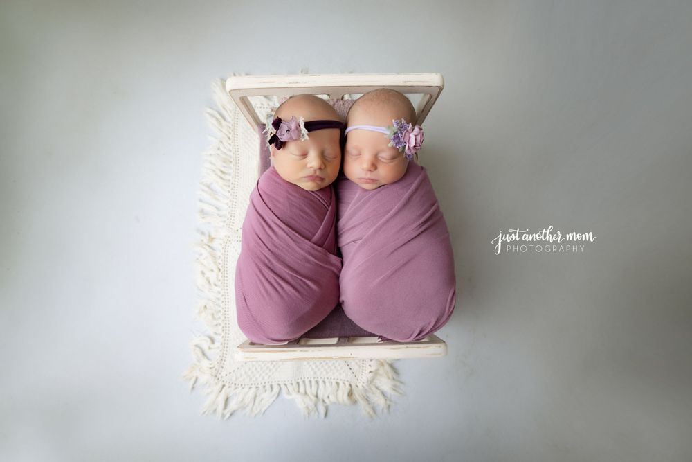 Two newborn twins swaddled in purple wraps lie side by side on a white textured blanket.