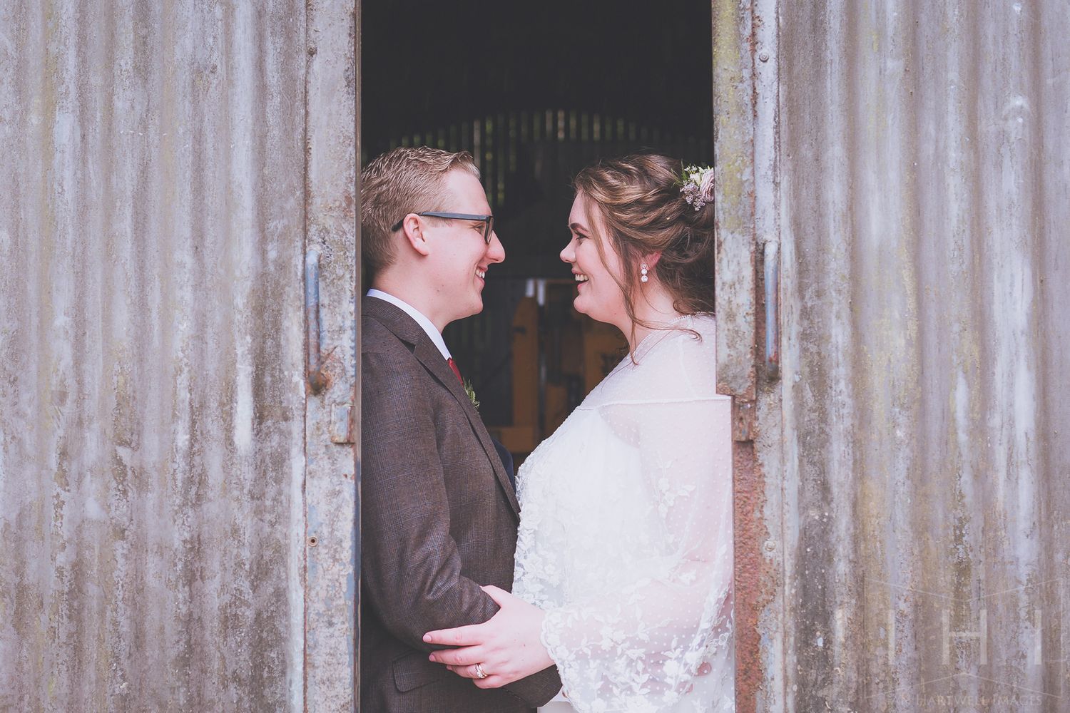 A bride and groom stand face to face, embracing each other and smiling. Framed by corrugated iron barn doors.