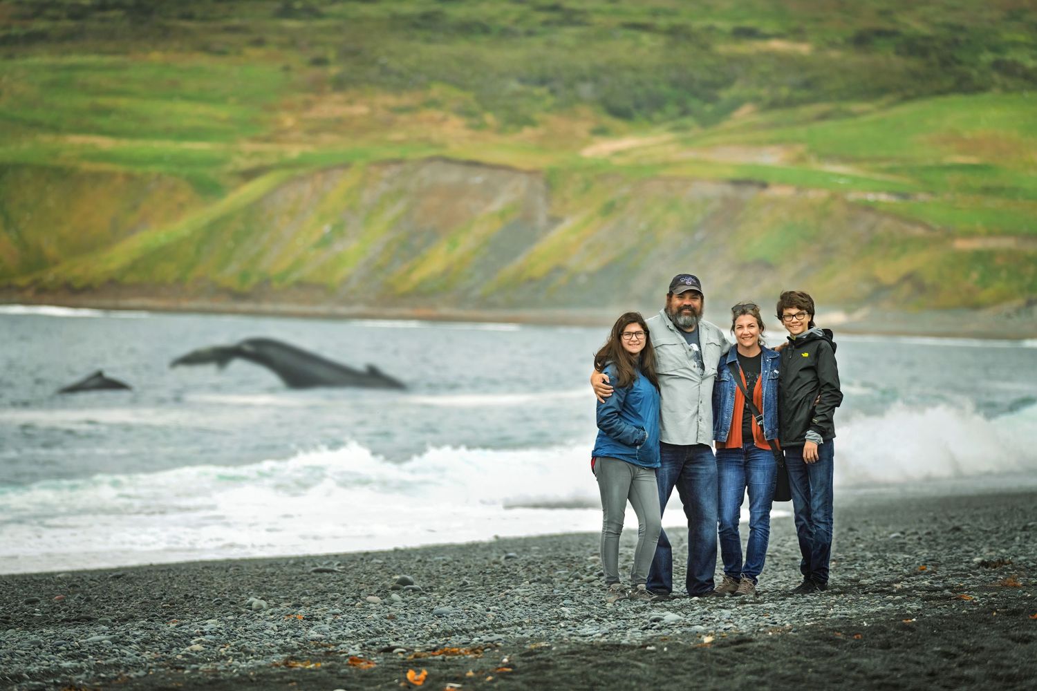 Group standing together on a rocky beach with crashing waves and green hills in the background.