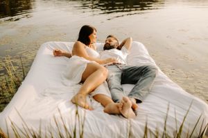 A couple relaxing together on a white mattress by a tranquil lake at sunset.