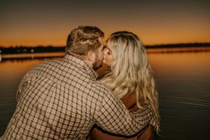 Intimate moment captured between couple against orange sunset over calm water.