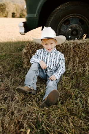 Young child in cowboy attire relaxes against hay bale near truck wheel in rustic outdoor setting.
