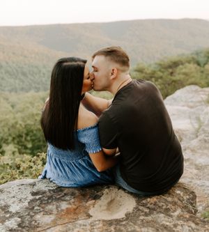 Couple in casual attire sharing tender moment while sitting on rocky mountaintop overlook.