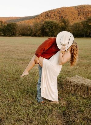 Couple in rustic attire sharing playful moment in field with mountains in background.