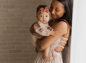 Mother holding baby in soft beige tones against textured brick wall background.
