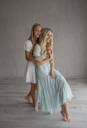 Two women in romantic vintage dresses embrace while posing elegantly against a minimalist grey backdrop.