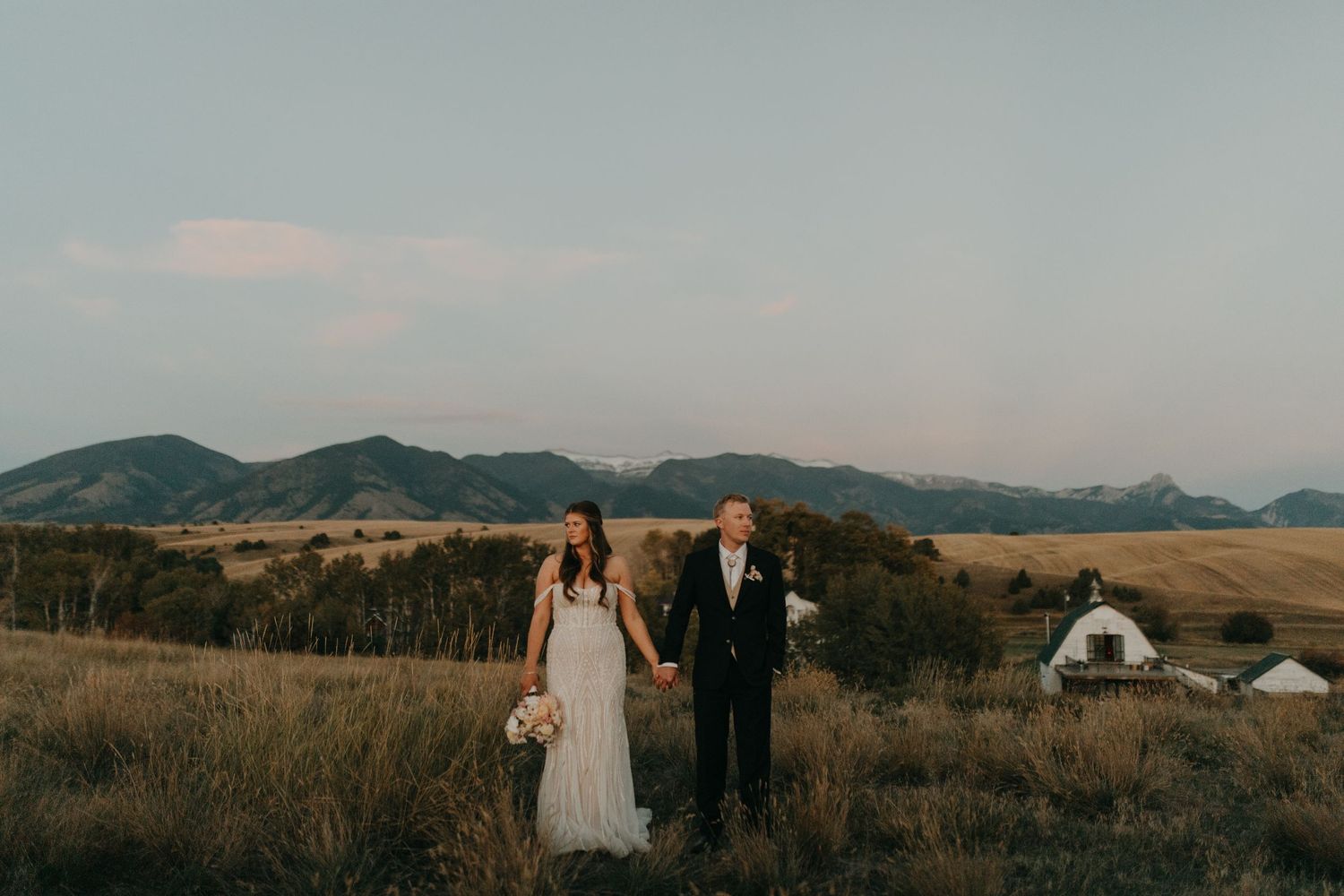 A wedding couple stands in a scenic mountain field with rustic buildings in the distance.