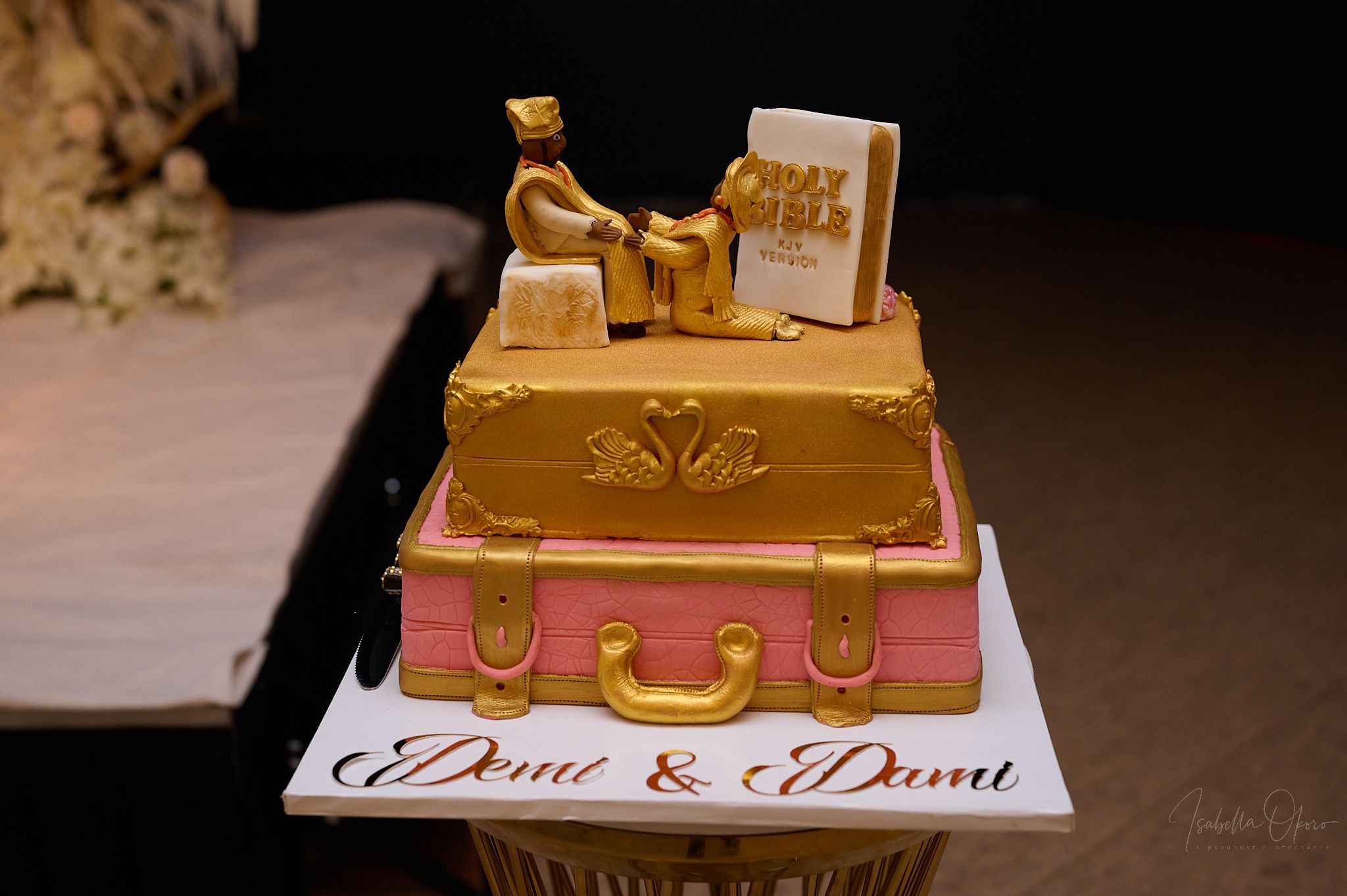 A gold-themed wedding cake with suitcase design and personalized topper reading Dami & Dami.