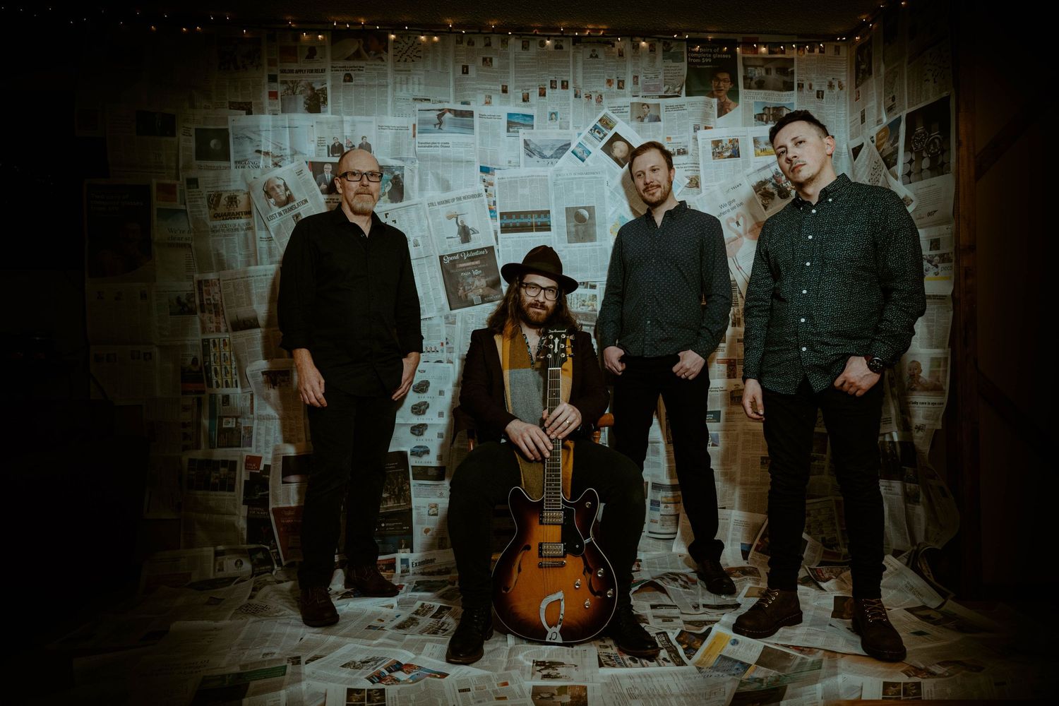 Musical group called John Hewitt and the New Americans poses together against a wall covered in papers for a promotional photo.