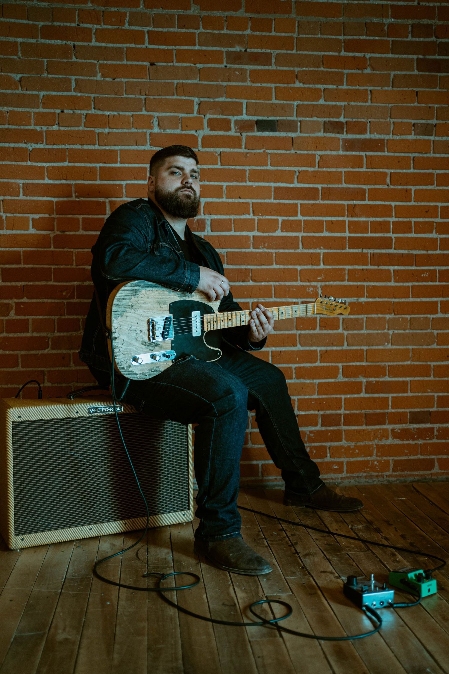 Dylan Farrell sits on an amp with a telecaster guitar against a brick wall backdrop.