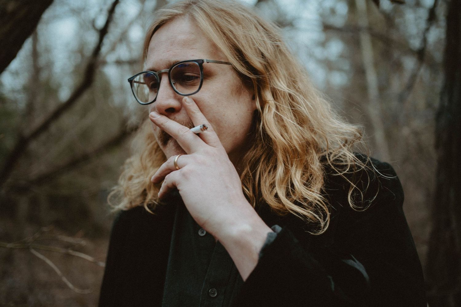 Person in glasses with long hair smoking in moody forest setting.