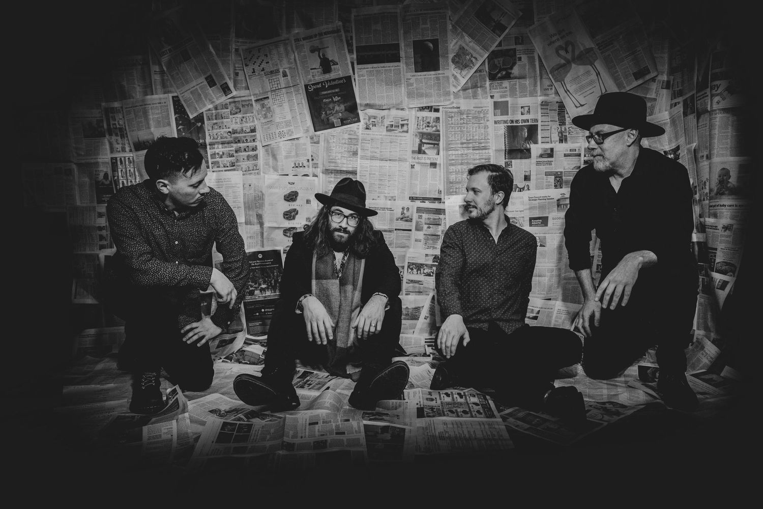 Musical group called John Hewitt and the New Americans sitting together against a wall covered in newspapers in black and white.