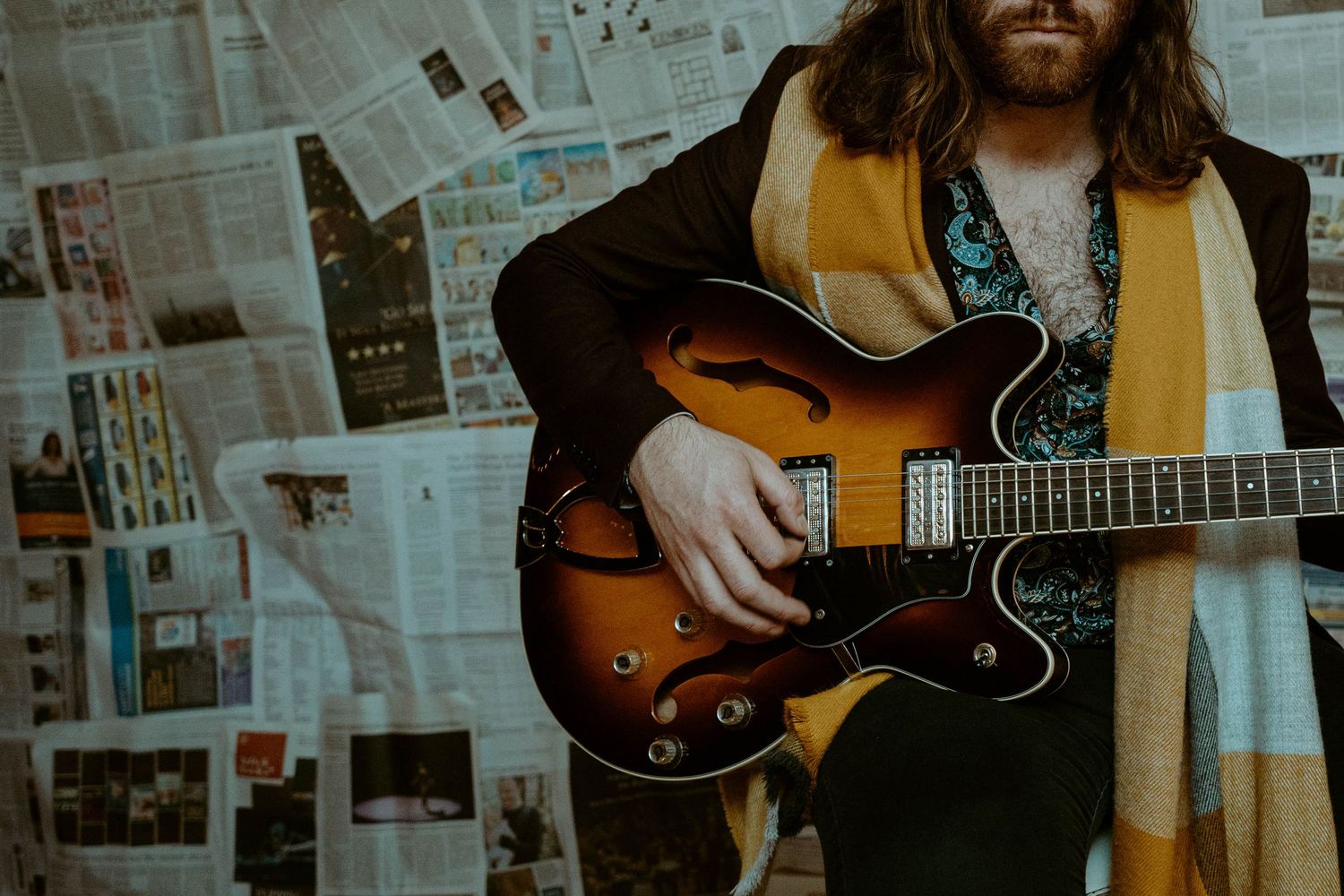 Close-up of a semi-hollow electric guitar being played by John Hewitt against a newspaper-covered wall.