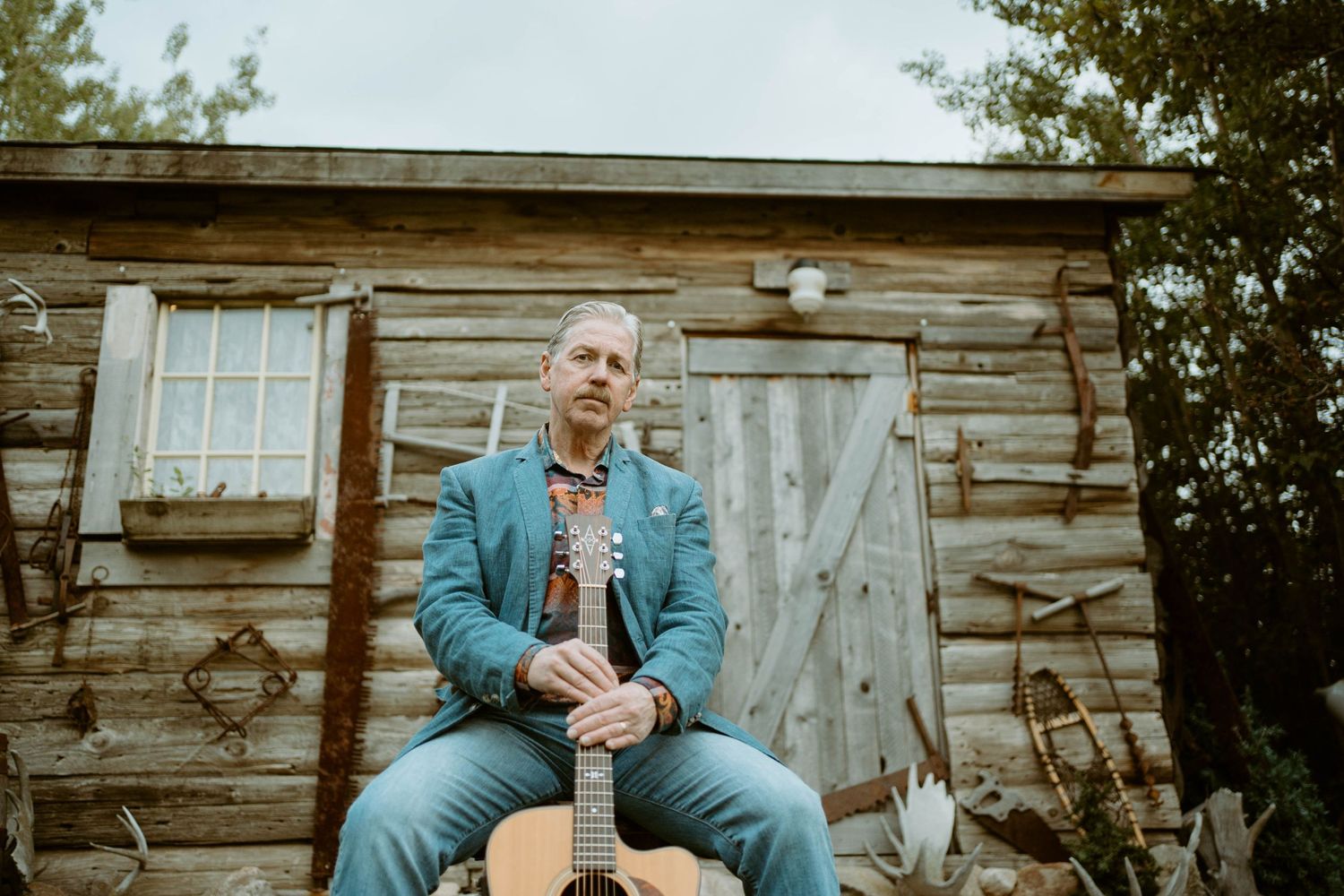 Tim Isberg in blue suit sits on wooden chair outside weathered cabin with guitar.