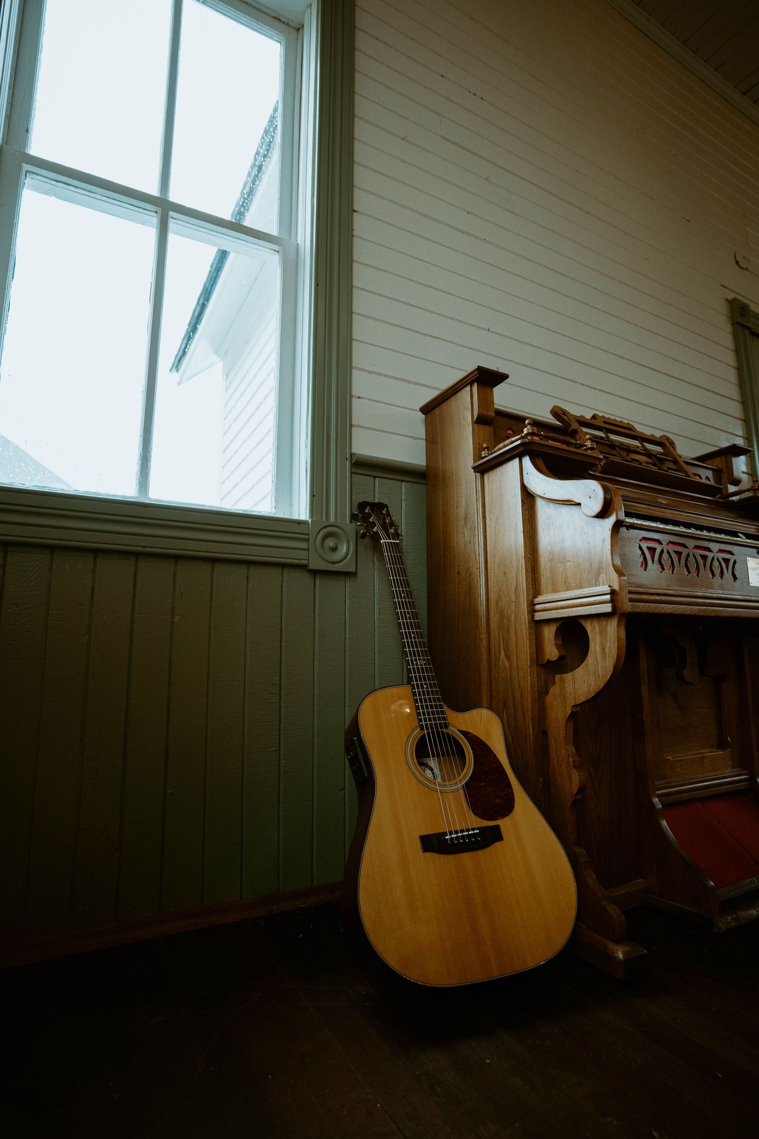 An acoustic guitar leans against a wooden piano near a sunlit window in a cozy room.
