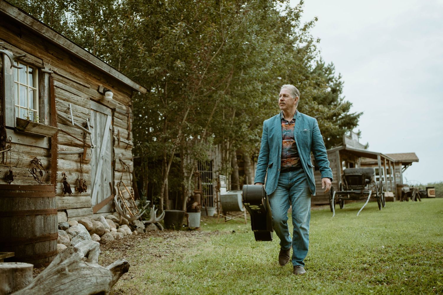 Tim Isberg in denim outfit walks alongside rustic wooden cabin in countryside setting.